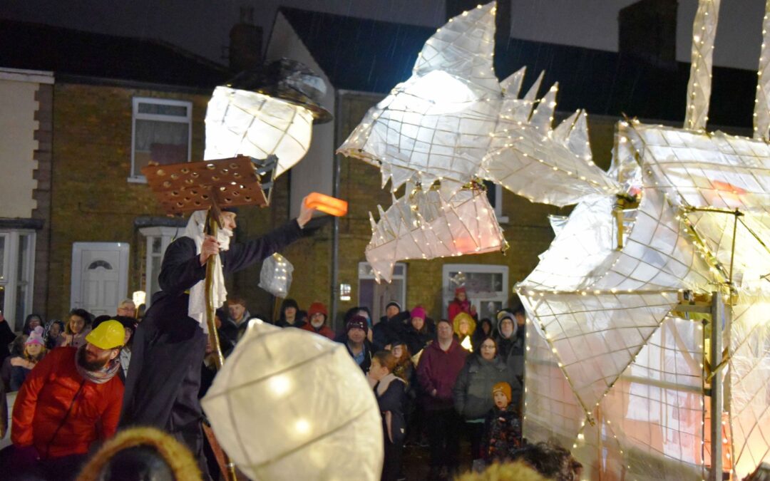 Large paper lantern structure in the shape of the "eye monster". The lantern is white and is lit from within with fairly lights. There is smoke coming from it's chimney on it's back. It's on a trailer. There are crowds around it on the street. It is being fed a 'red brick' lantern.