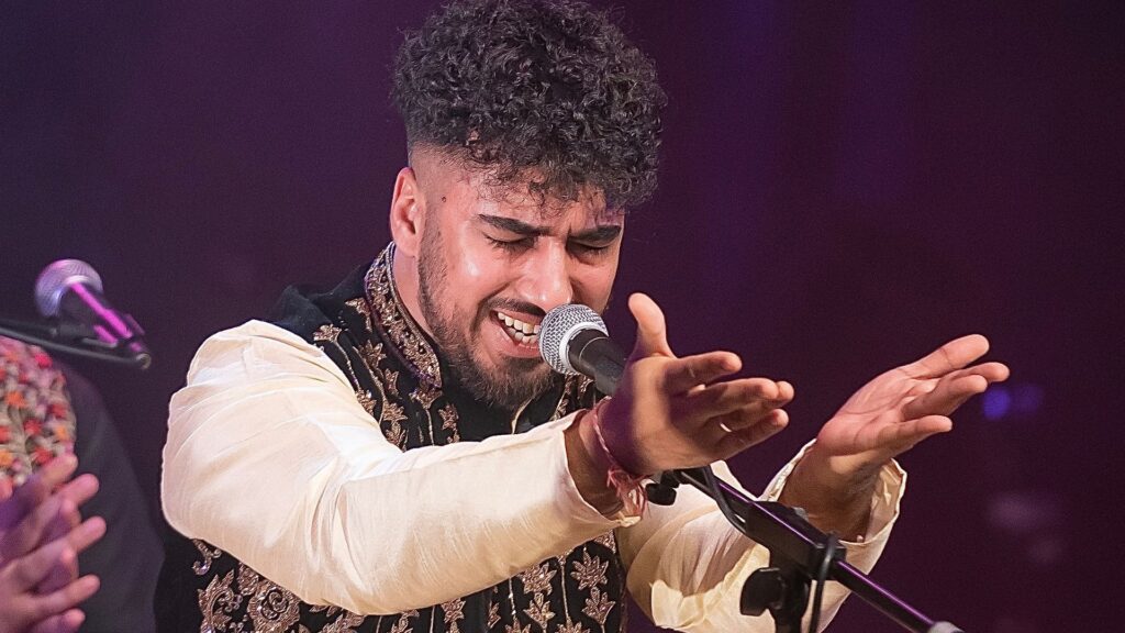 chand ali khan singing into the microphone. His eyes are closed and his hands are outstretched, palms upwards. He is wearing traditional shalwar kameez.