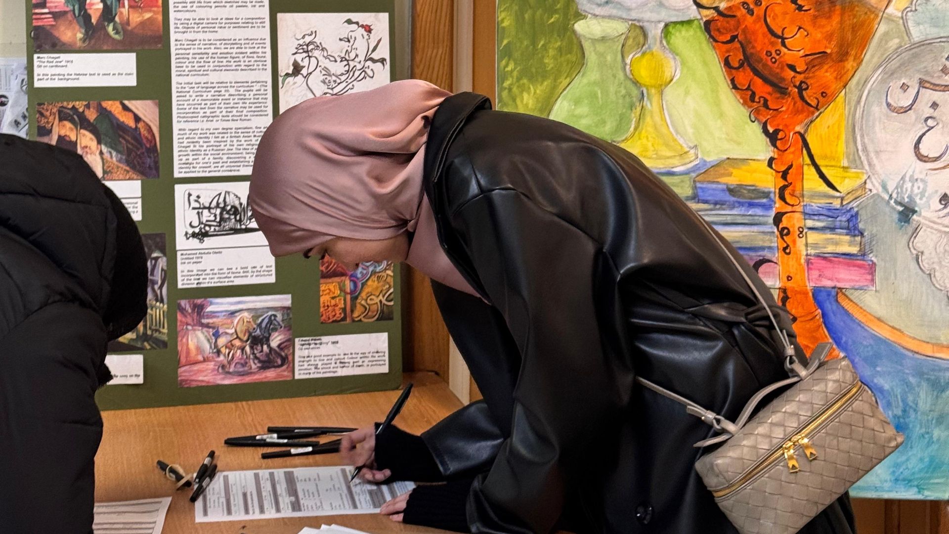 A photo of a young woman in a dusky pink silk hijab and black coat, leaning over as she fills in a form. Behind her, vibrant artwork with urdu script and curved shapes in orange, yellow, and green.